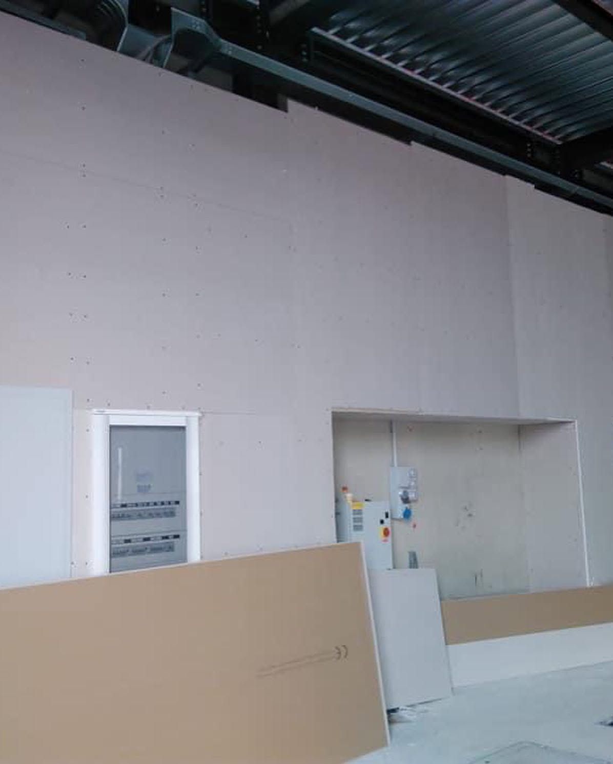 Interno industriale con un'ampia apertura nella parete, una porta e pannelli di costruzione in primo piano.
