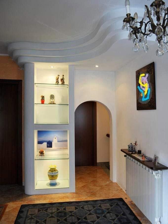 Luminoso corridoio con mensole illuminate a incasso, porta ad arco, lampadario e quadri incorniciati alle pareti.