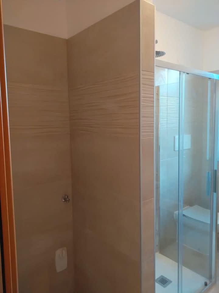 Bagno piastrellato di beige con box doccia in vetro e WC a vista in un angolo.