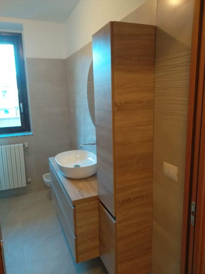 Bagno moderno con mobiletto in legno, lavabo a ciotola bianco, finestra e termoarredo.