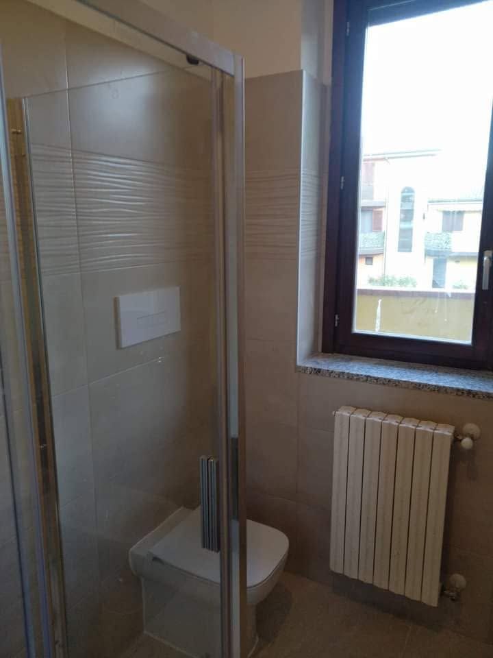 Piccolo bagno con doccia in vetro, WC bianco e finestra sopra il termosifone.