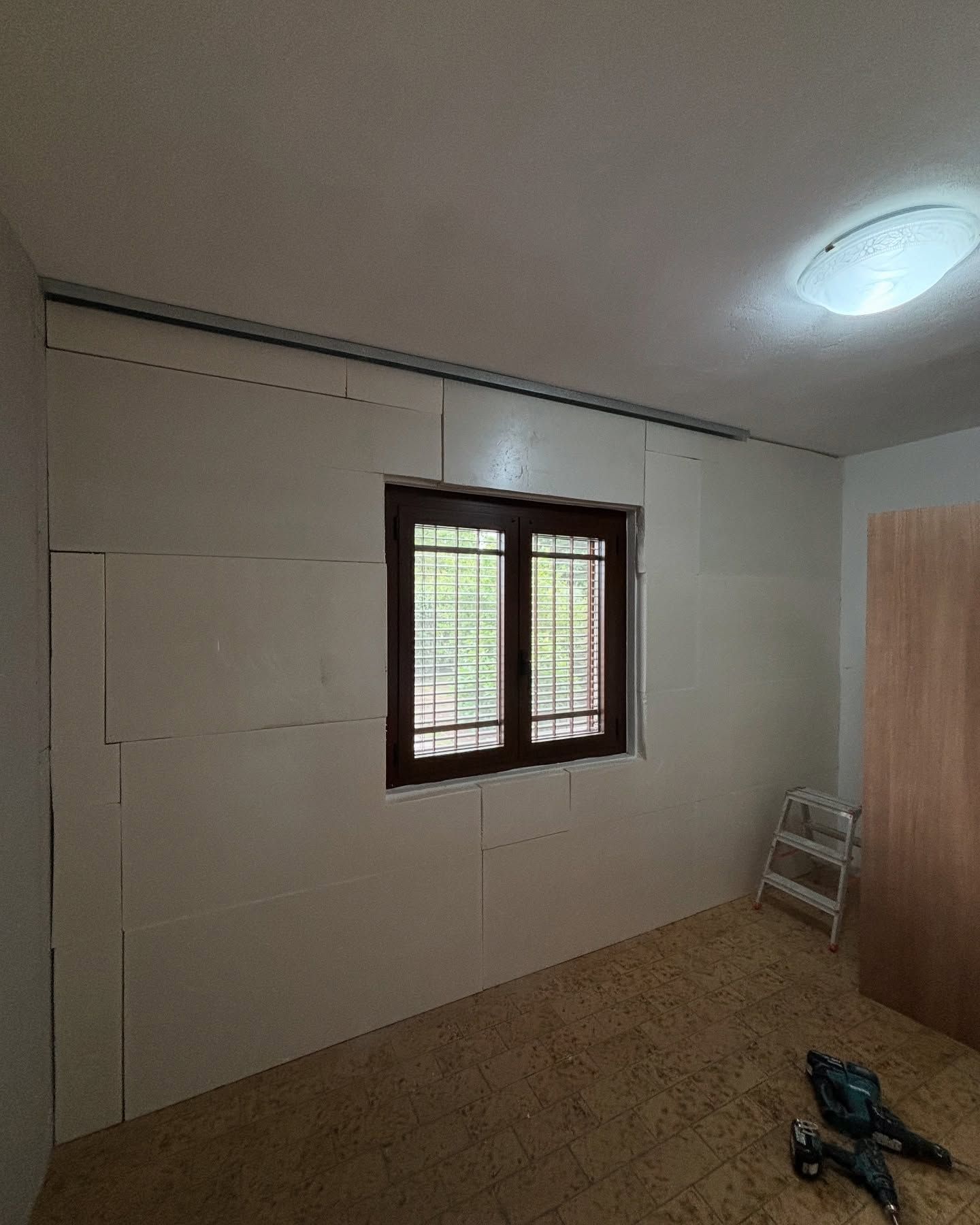 Stanza vuota con pareti rivestite di pannelli bianchi, una finestra, una lampada a soffitto e una scala con attrezzi sul pavimento.