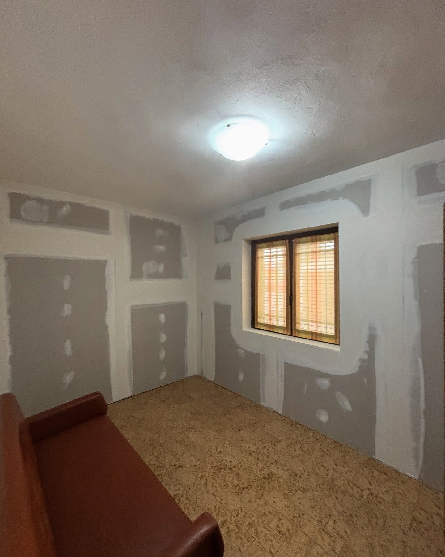 Stanza vuota in fase di ristrutturazione con toppe di cartongesso grigie, una finestra e una lampada a soffitto.