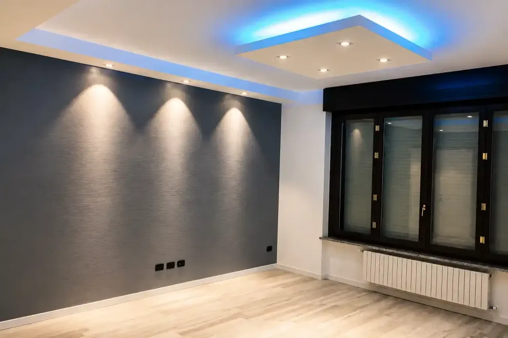 Stanza moderna vuota con parete d'accento grigia, faretti a incasso, luce blu sul soffitto e un grande termosifone a panca sotto la finestra.