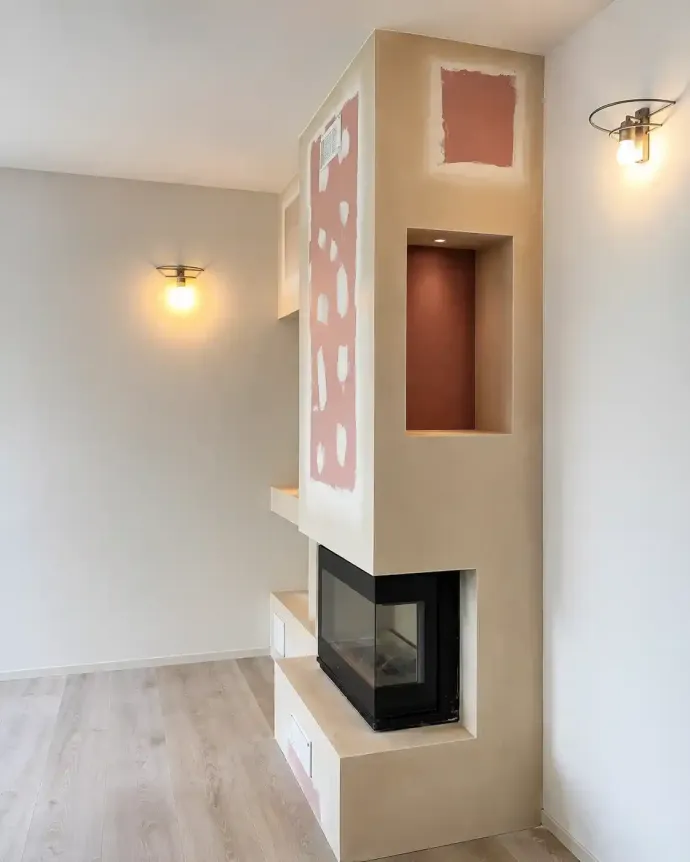 Camera moderna con camino ad angolo beige, pannello decorativo rosa, applique a muro e pavimento in legno chiaro.