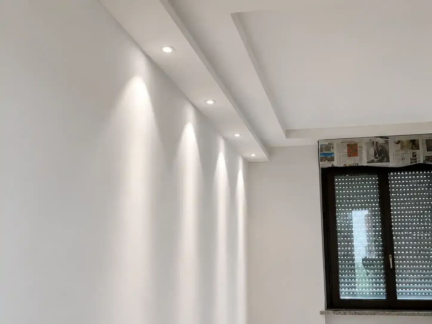 Stanza con interni bianchi, faretti a incasso sul soffitto e una finestra con tende scure.