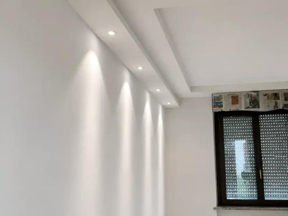 Stanza con interni bianchi, faretti a incasso sul soffitto e una finestra con tende scure.