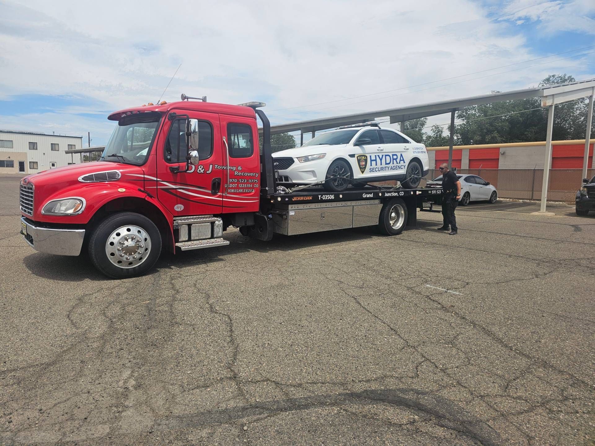 Red flatbed tow truck carrying a white vehicle in a parking lot under a cloudy sky