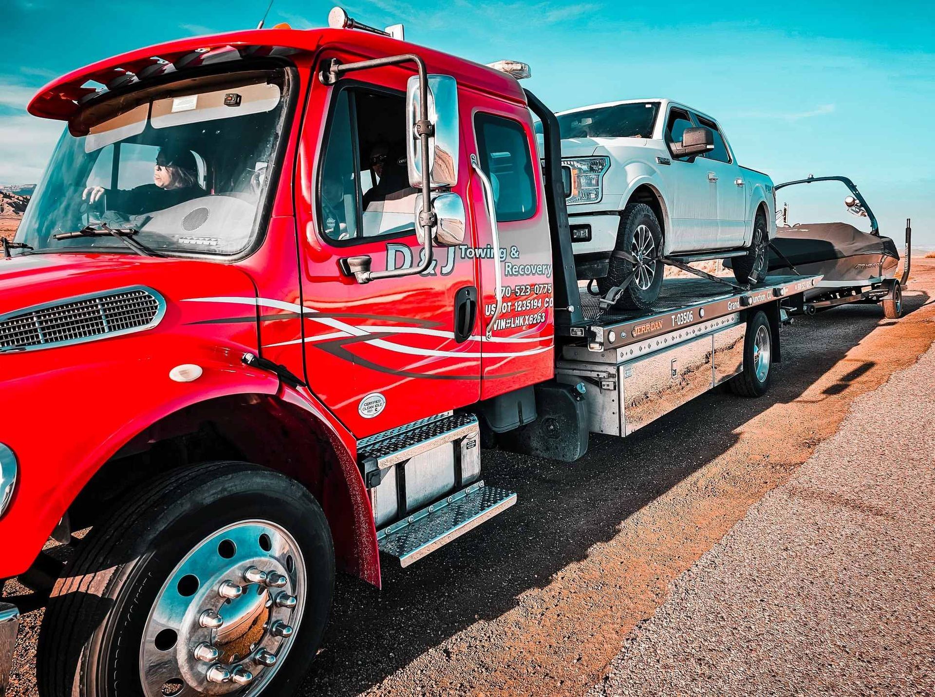 Red tow truck hauling a silver car on a flatbed along a roadside under a blue sky