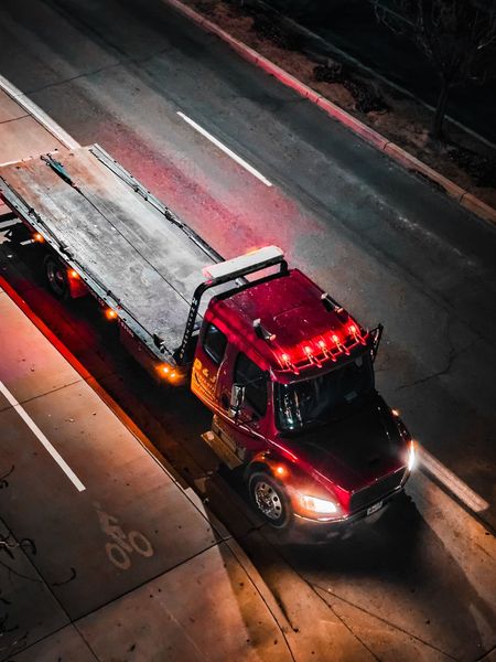 Red tow truck driving at night on a city street, headlights on, with flashing red lights on top.