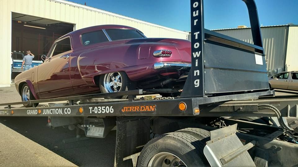 Maroon classic car strapped on a flatbed tow truck, parked outdoors in bright sunlight.