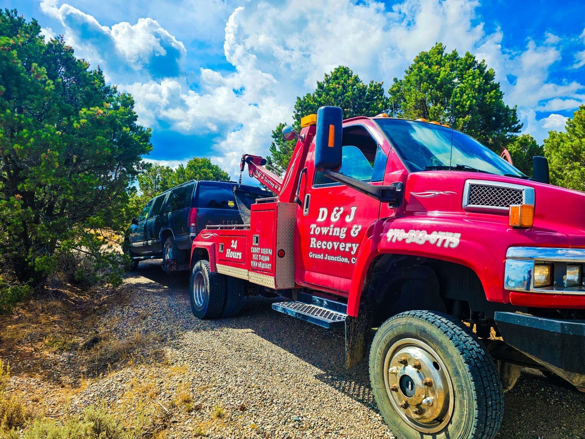 Red tow truck on gravel road with green trees and blue sky, towing a black vehicle
