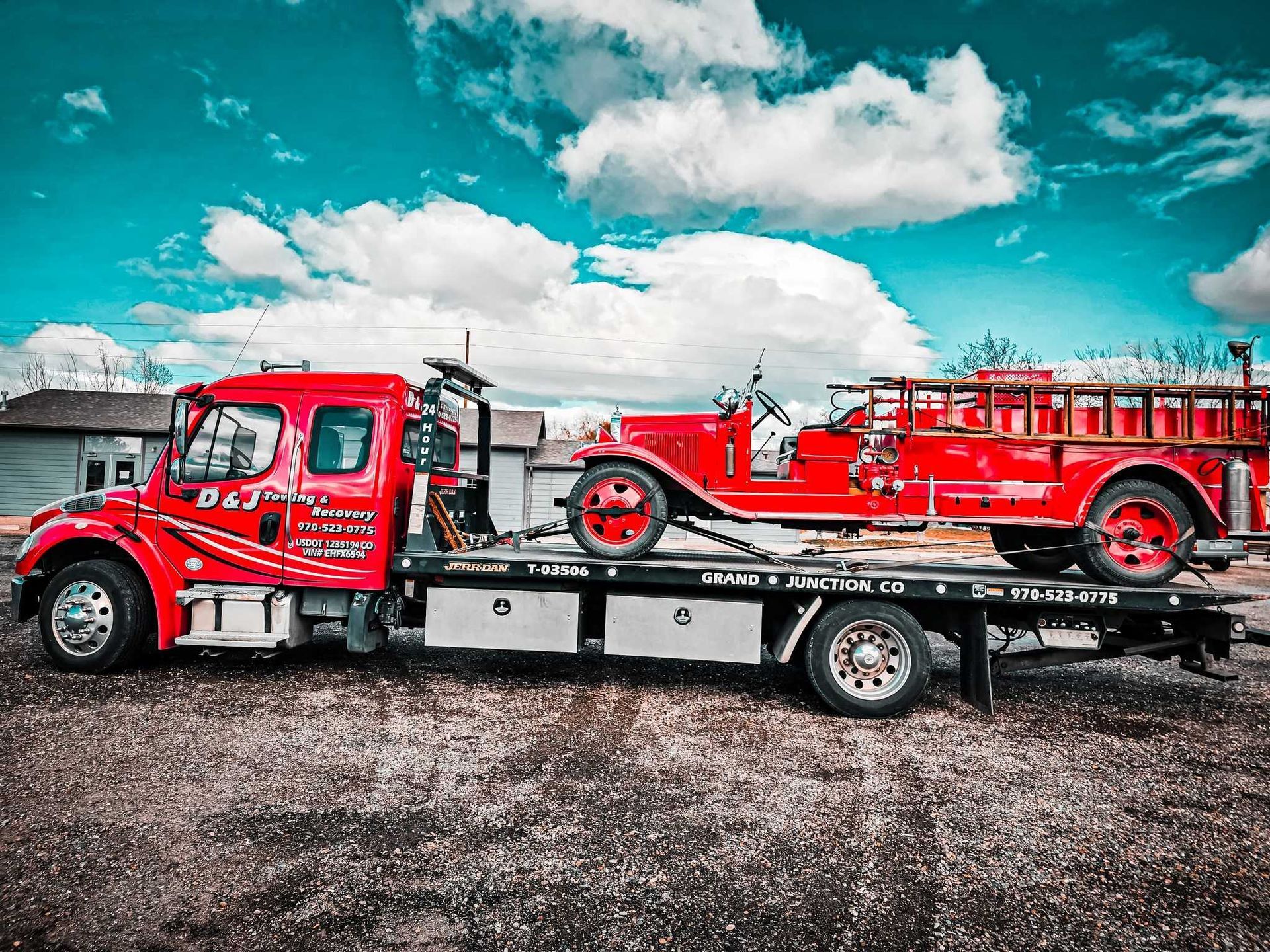 Red tow truck carrying a red vintage fire truck on a flatbed trailer under a cloudy sky