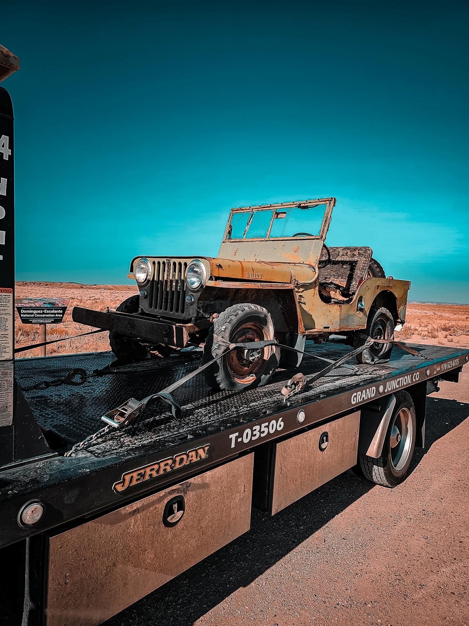Rusty old jeep on a flatbed trailer in a desert under a bright blue sky