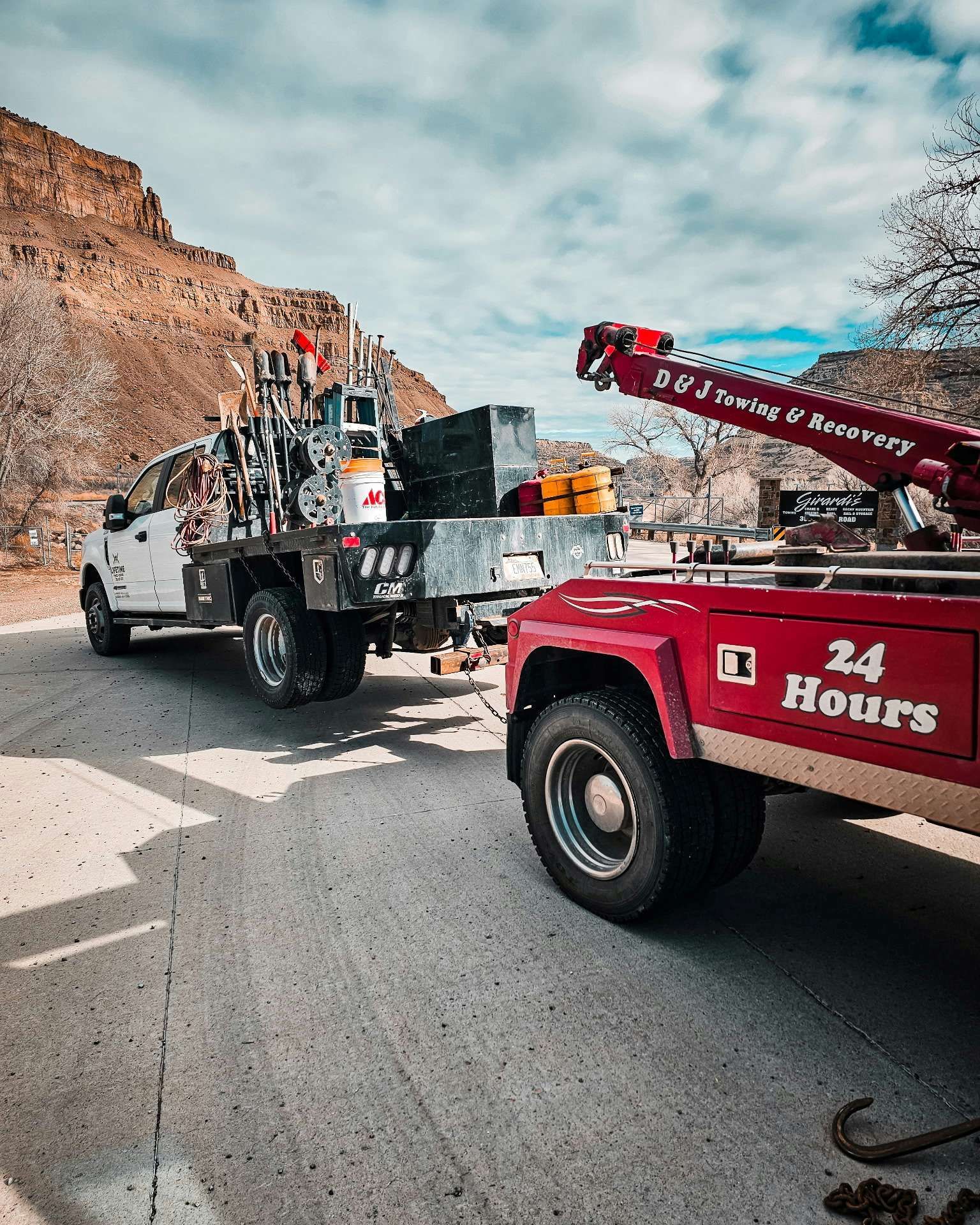Two tow trucks on a mountain road, one red and one white, against a rocky cliff and cloudy sky