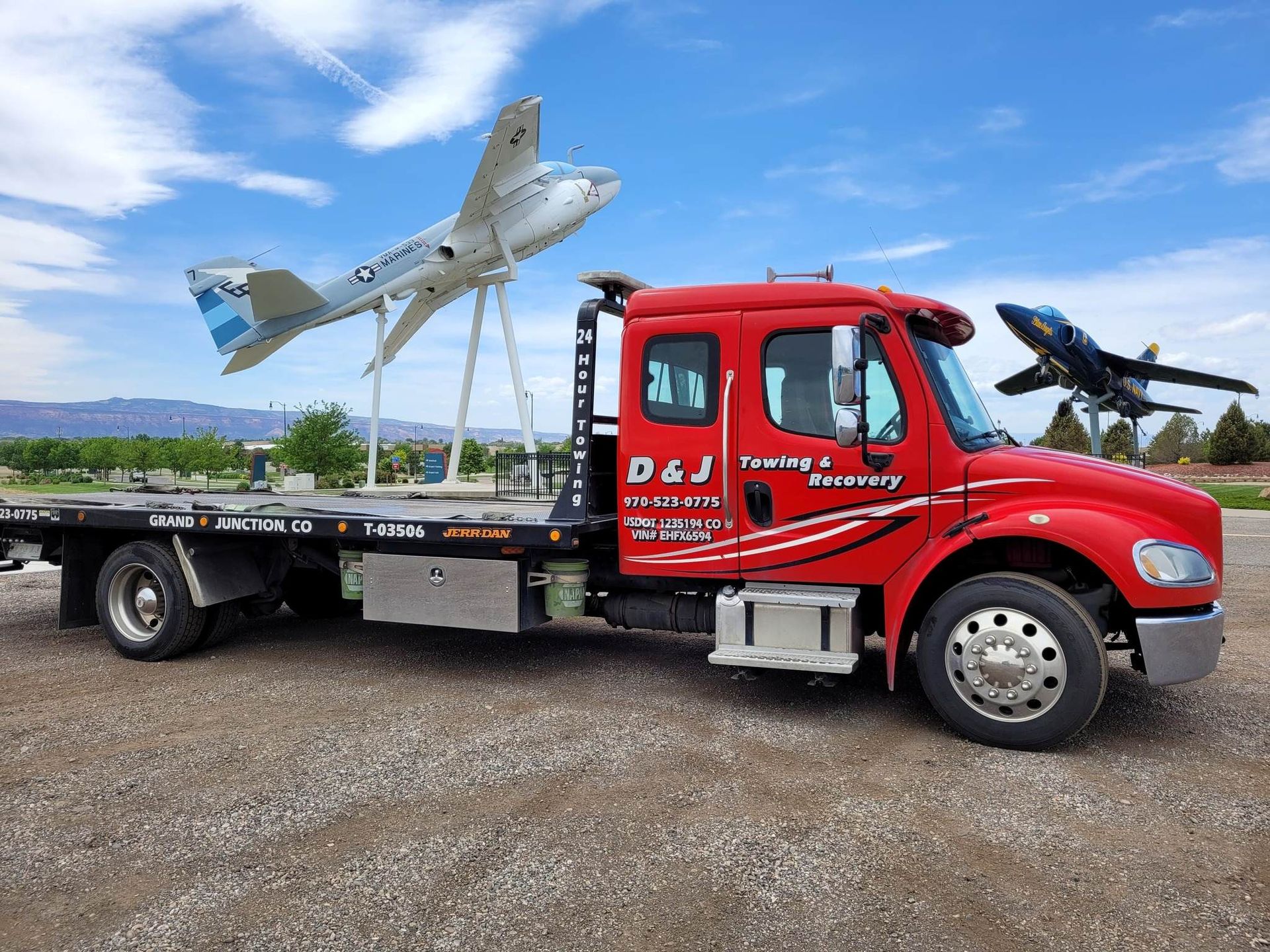 Red flatbed tow truck carrying a small airplane and a drone in a gravel lot under a blue sky