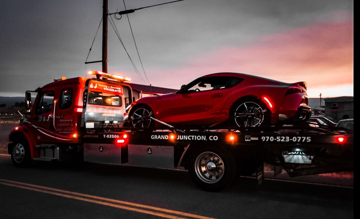 Red sports car loaded on a tow truck at dusk, with orange lights and a city skyline in the background