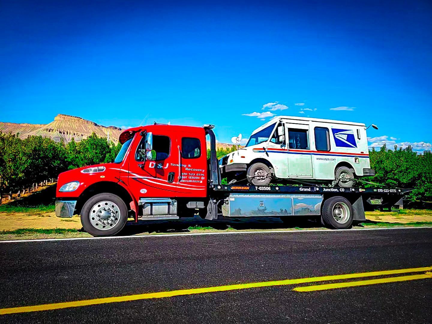 Red tow truck carrying a white SUV on a flatbed beside a road under a bright blue sky.
