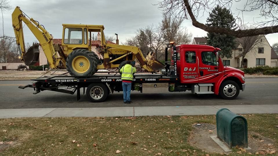 Red flatbed truck carrying a yellow backhoe, with a worker standing beside it on a suburban street.