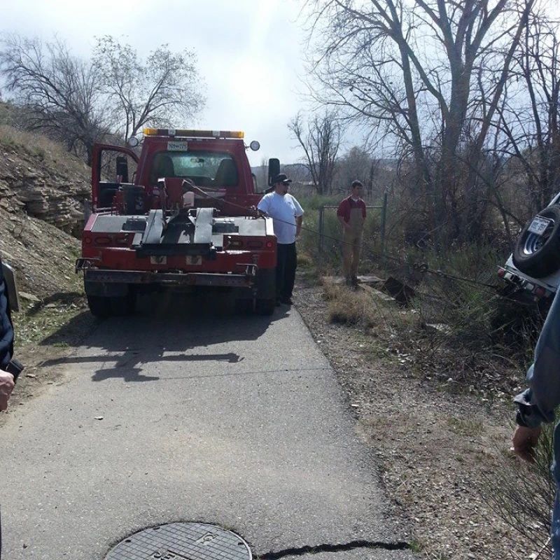 Red tow truck blocking a narrow road beside two people in a sparse rural area