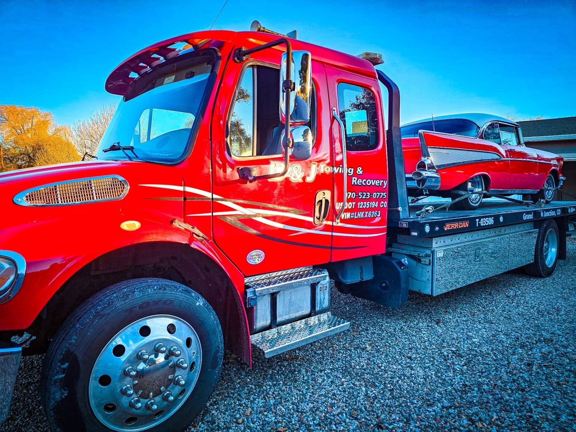 Red tow truck carrying a red classic car on a flatbed in a gravel lot