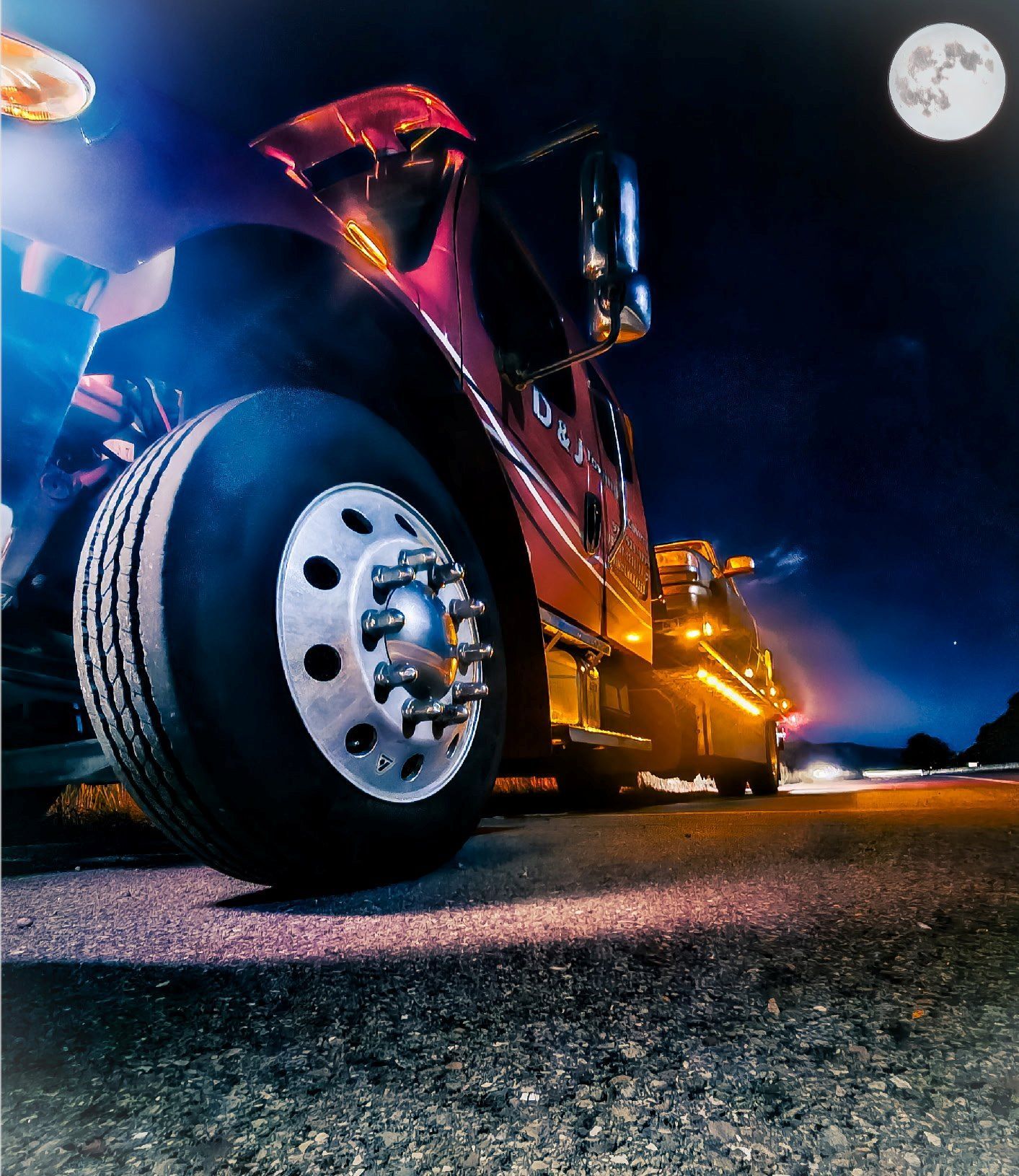 Low-angle view of a shiny red semi truck with bright lights driving at night under a full moon