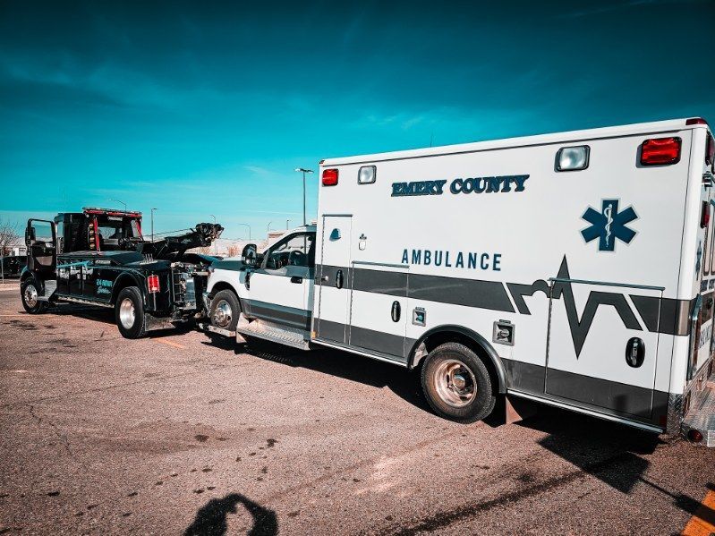 Ambulance towed by a truck on a rural road under a bright blue sky