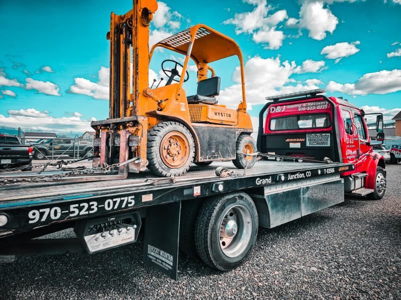 Orange forklift on a black flatbed tow truck in an industrial lot under a blue sky.