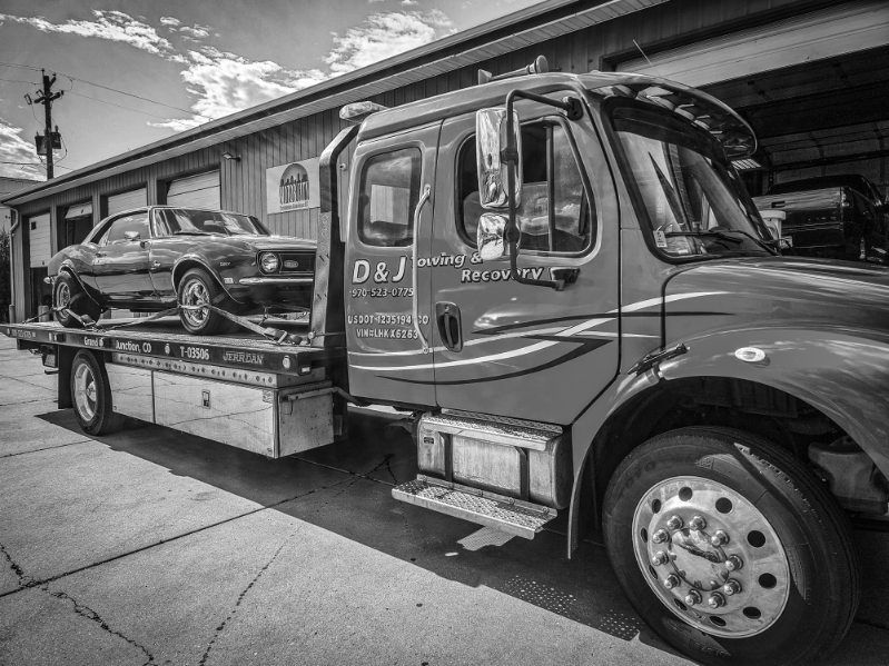 Tow truck hauling a black car on a flatbed beside a building under an overpass