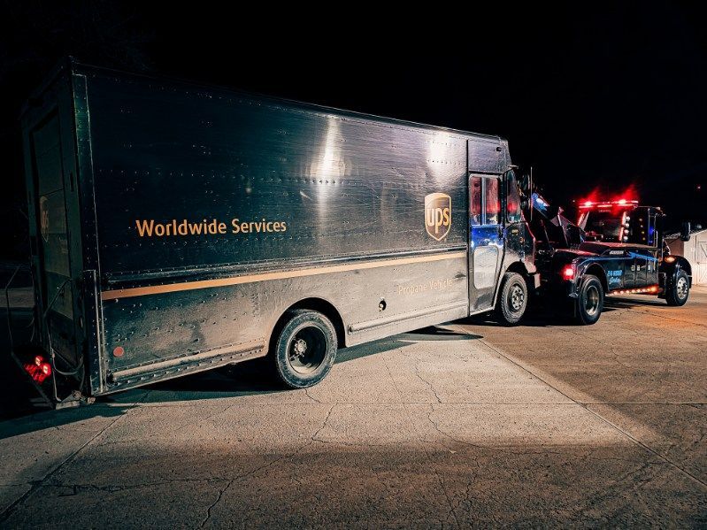 Dark box truck hitched to a blue tow truck at night, with red lights flashing on a roadside.