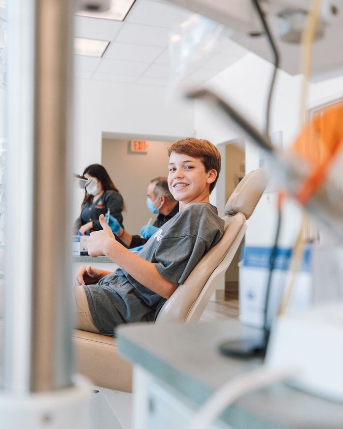 A young boy is sitting in a dental chair giving a thumbs up.