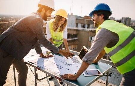 Construction team reviewing blueprints at a job site, wearing hard hats and high-visibility vests.