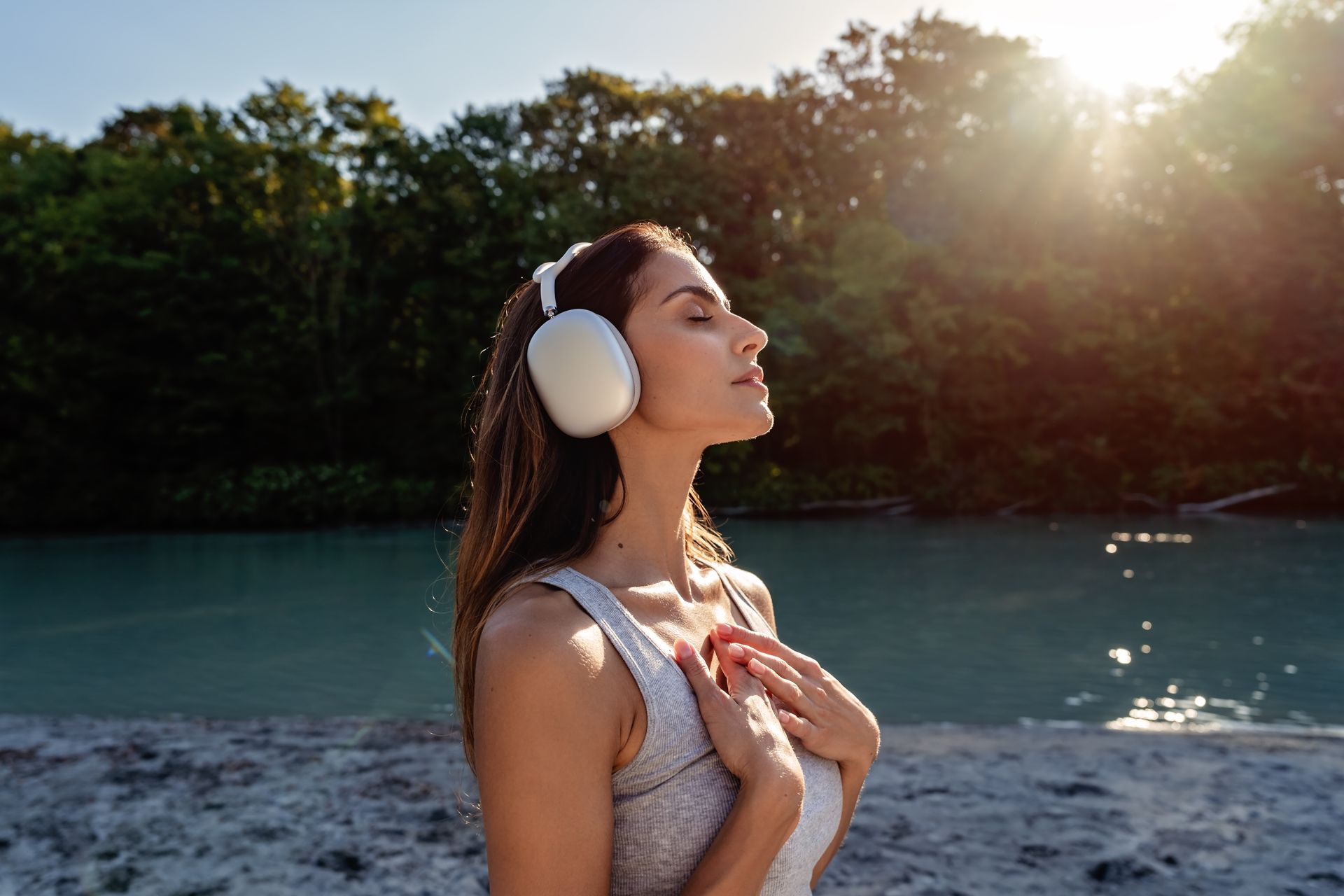 Woman wearing white headphones stands by a lake at sunset with eyes closed and hands on chest.