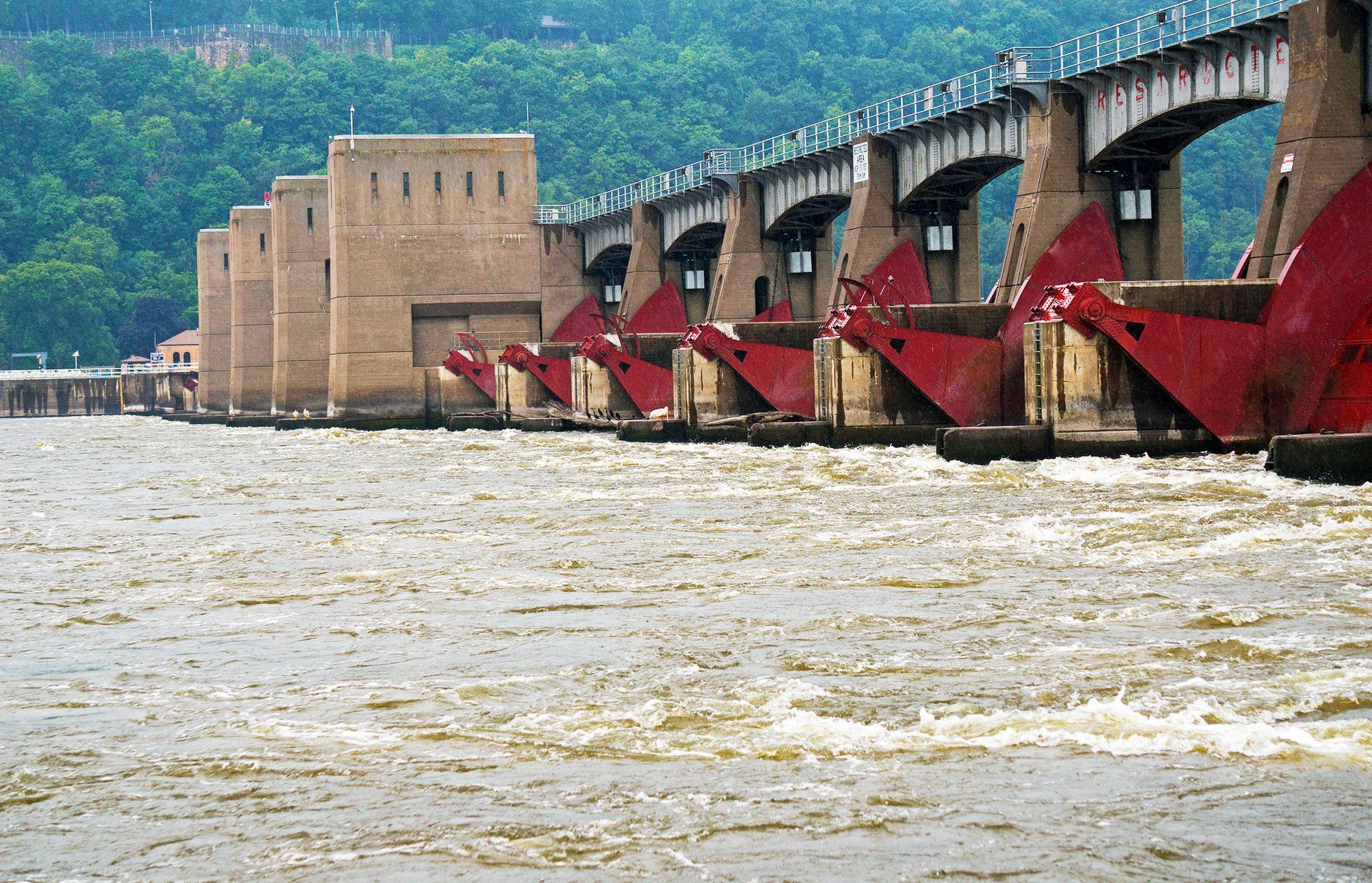 A series of red floodgates at a dam on a wide, churning river, set against a backdrop of a lush green hillside.