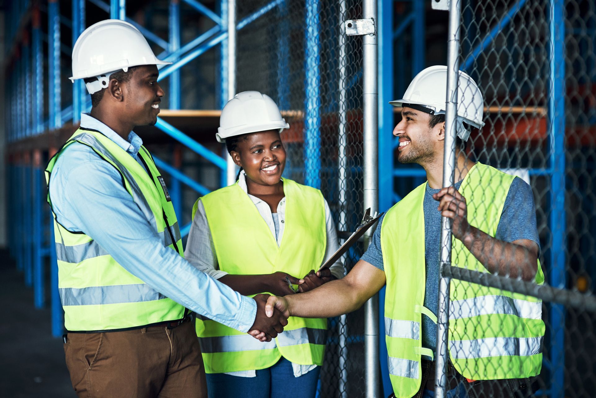 Three workers in a warehouse shake hands, wearing hard hats and safety vests.
