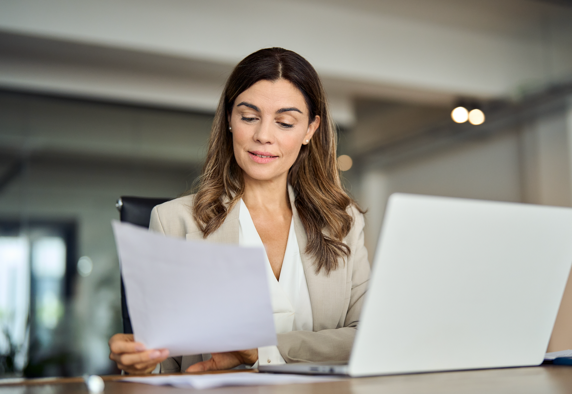 A woman is sitting at a desk with a laptop and a piece of paper.