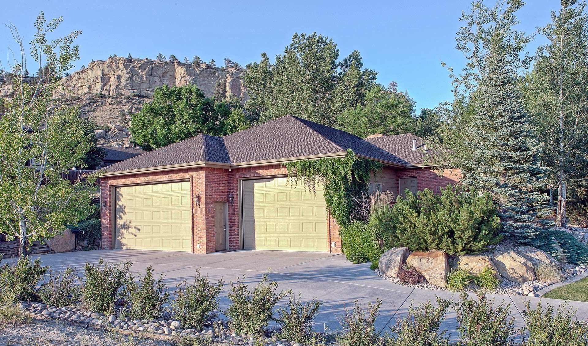 Two-car garage with tan doors and brick trim, set among trees beneath rocky cliffs.