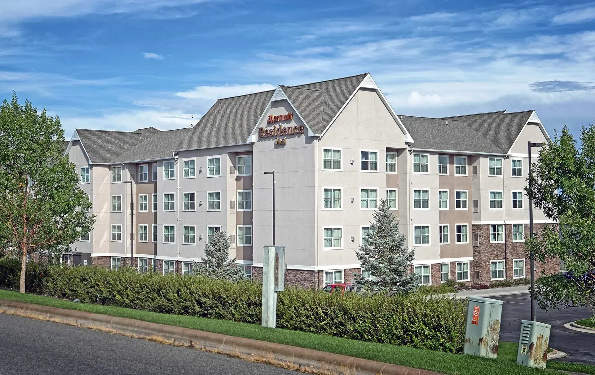 Three-story cream apartment building with gray roofs beside a road and trees under a blue sky