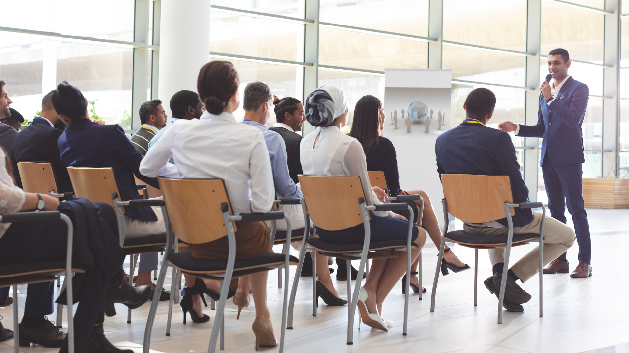Audience seated in a bright conference room watching a speaker present at the front