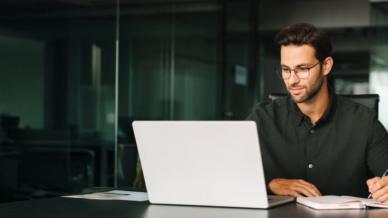 Man working at a laptop in a modern office, with a notebook beside him.