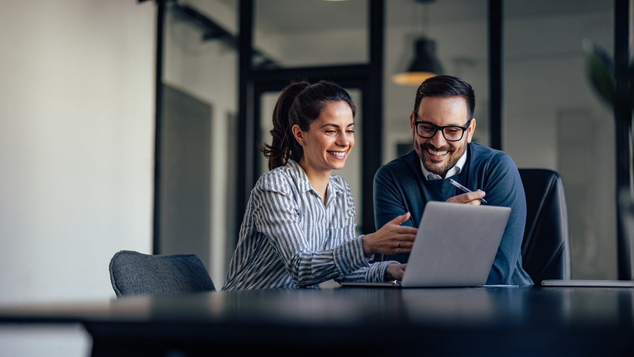 Two coworkers smiling and reviewing a laptop together in a modern office