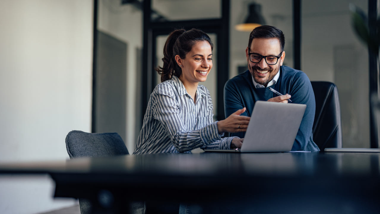 Two coworkers smiling and reviewing a laptop together in a modern office