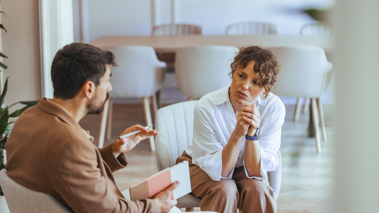 Two people talking in a bright office, one holding papers and the other listening thoughtfully.