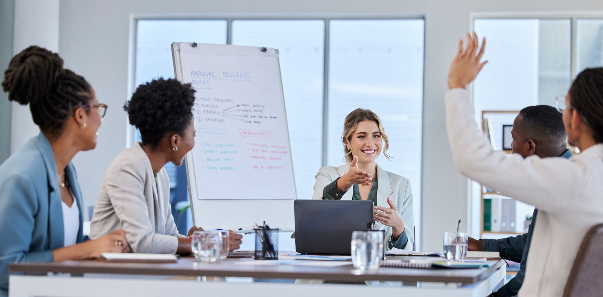 Two people in a meeting room, one gesturing while speaking across a laptop.