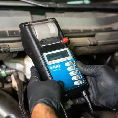 Gloved hands holding a blue handheld diagnostic meter inside a car engine bay