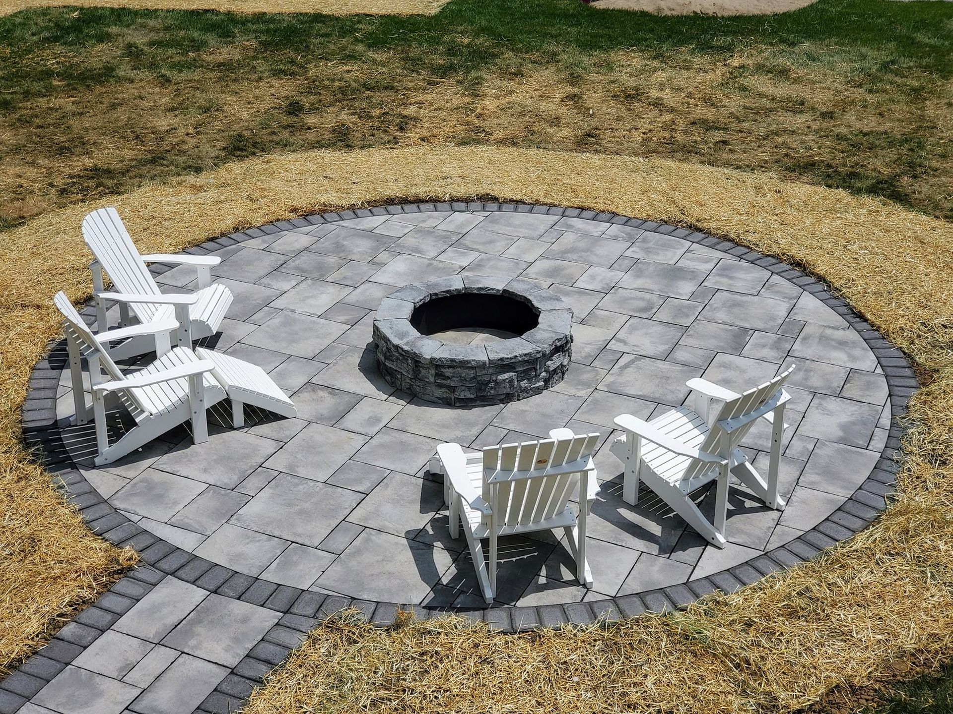 Circular paver patio setup with a central block fire pit and white adirondack chairs.