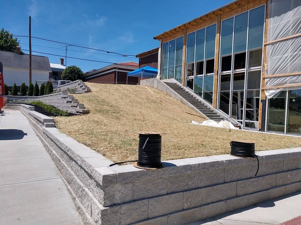 Large gray block retaining wall framing a grassy hill next to a modern commercial building with floor-to-ceiling windows