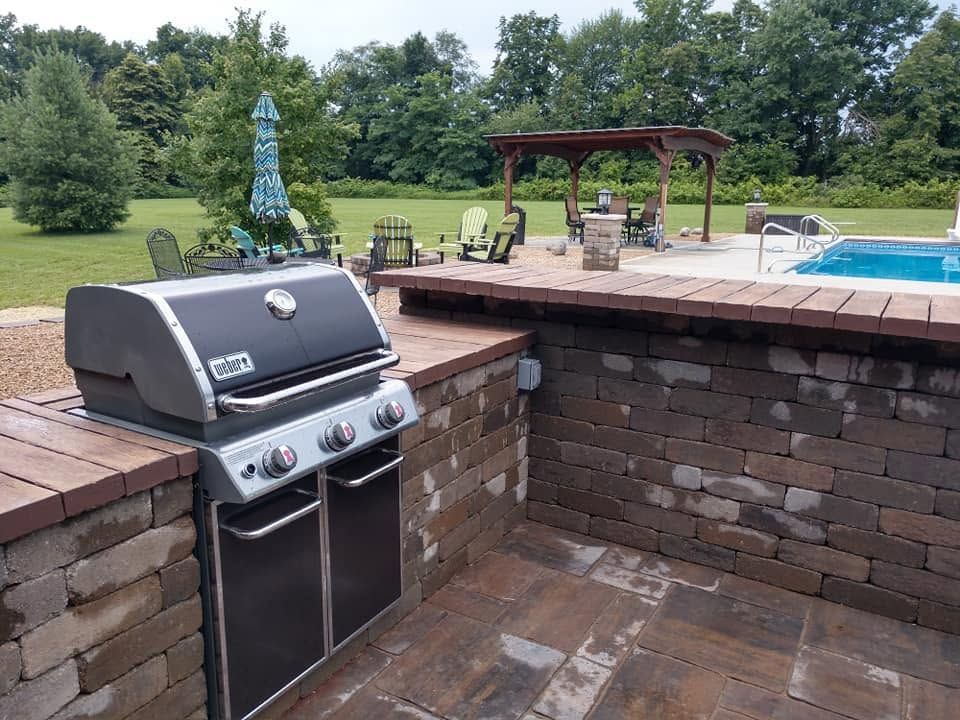 Brick outdoor kitchen with stainless grill beside a pool and patio, overlooking a grassy yard and trees