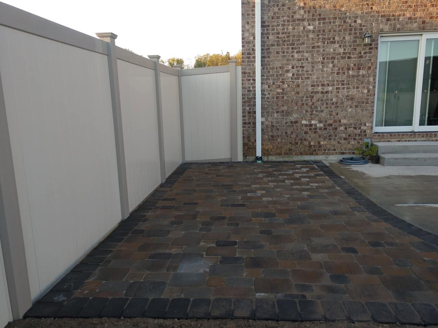 Dark block paver patio walkway neatly enclosed between a white privacy fence and a brick building wall
