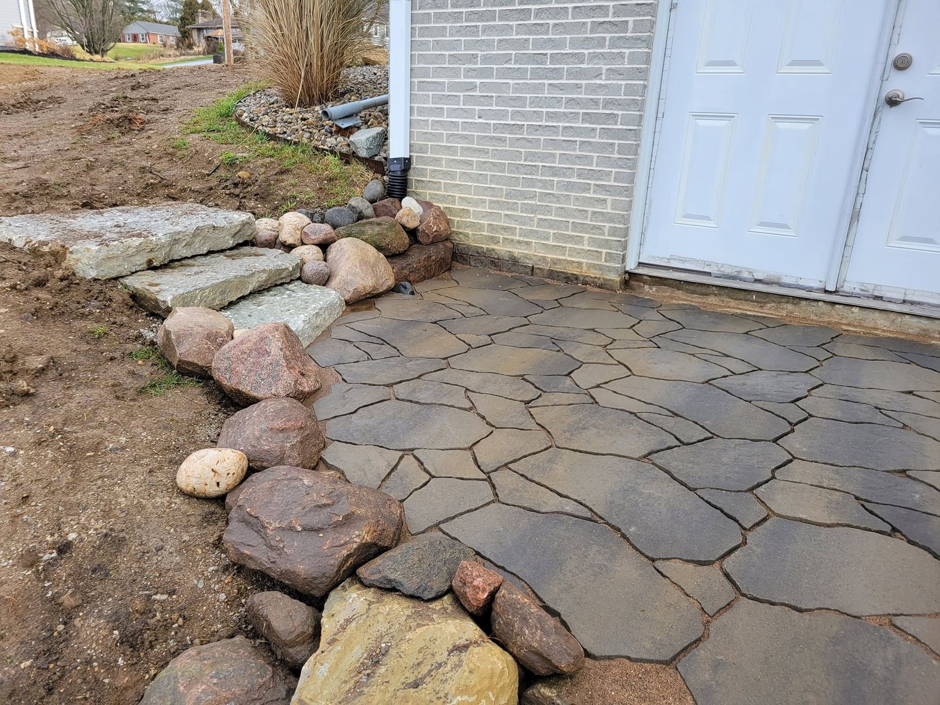Stone-paved patio beside a white door, bordered by large rocks and a dirt garden bed
